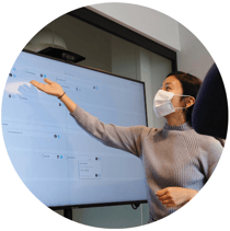 woman-at-work-pointing-to-screen-in-mask-and-grey-sweater